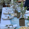 A table setting viewed from the head of the table shows plates with white napkins, silver napkin rings and sprigs of fresh rosemary. Four olive trees act as a centerpiece down the middle of the table.
